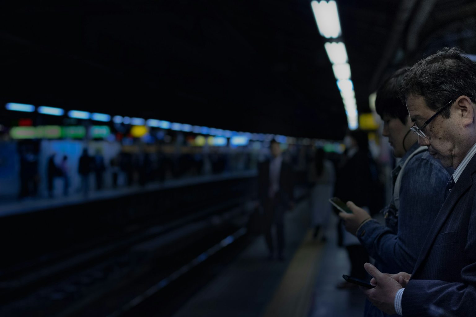 駅のホームで電車を待ちながらスマートフォンを見る会社員たち