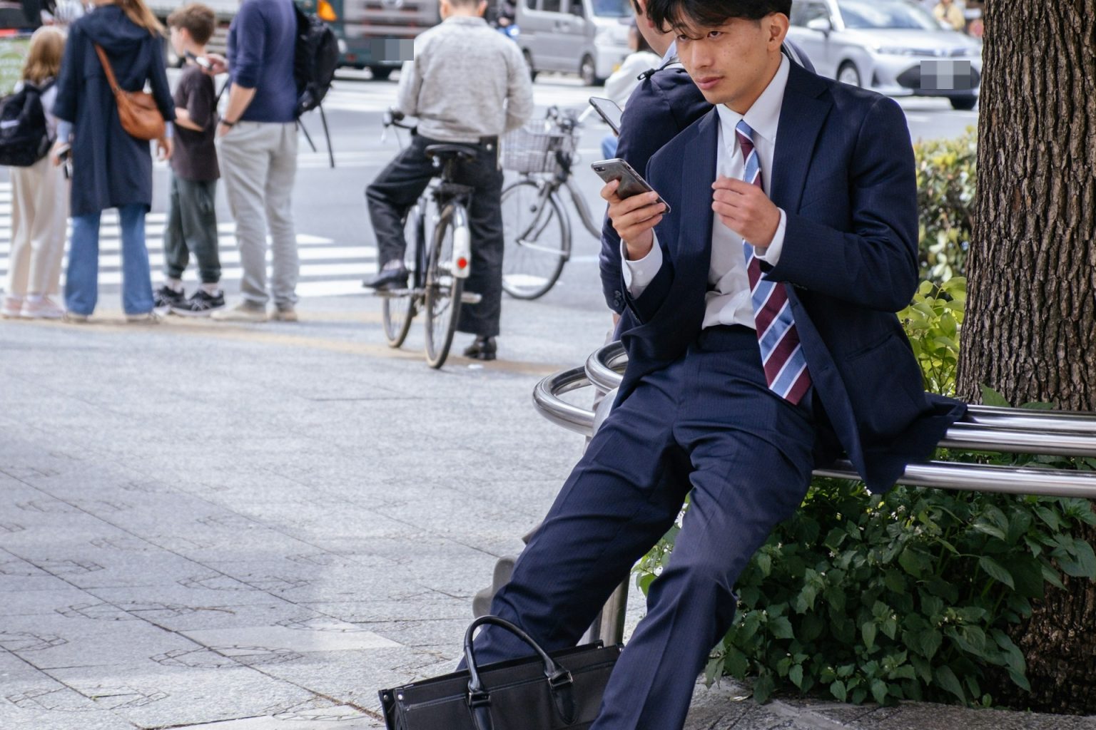 街路樹の下の手すりに腰掛け、スマートフォンを見るスーツの若い男性